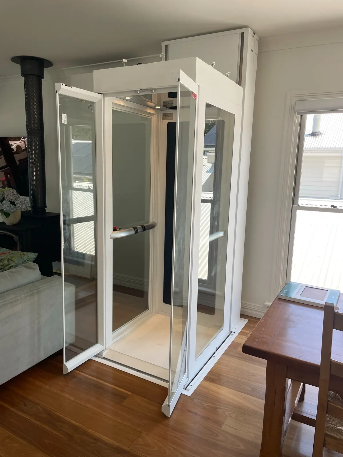 Lift rumah Ascenda dengan kabin kaca dan frame putih, pintu terbuka di ruang keluarga, solusi akses antar lantai tanpa mengorbankan area interior.