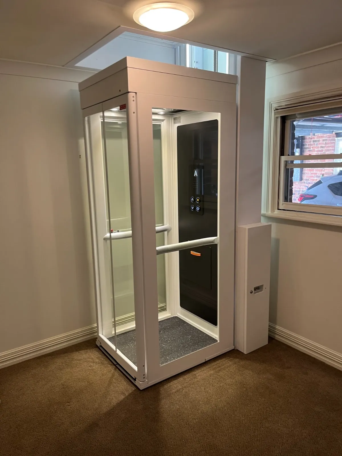 Alt text: Instalasi lift rumah Ascenda di area koridor, kabin kaca dengan panel kontrol di dalam, pemasangan rapi untuk rumah 2 lantai dengan ruang terbatas.