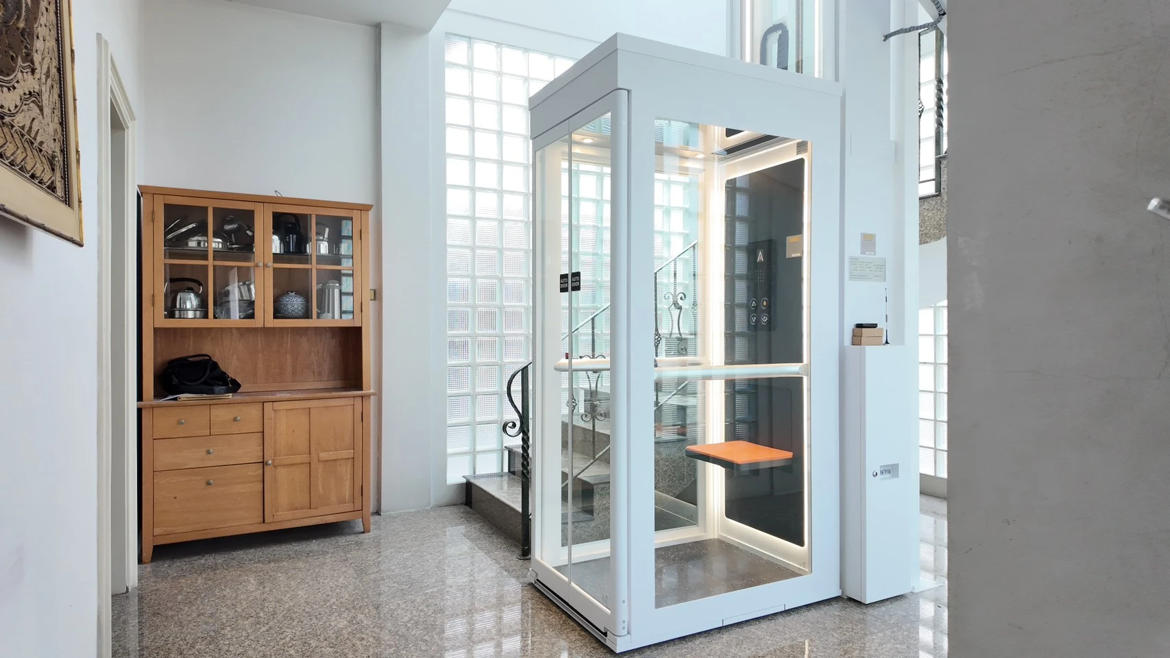 Lift kaca Ascenda by Cibes terpasang di area tangga dengan dinding glass block, tampilan sudut yang terang dan rapi untuk interior rumah