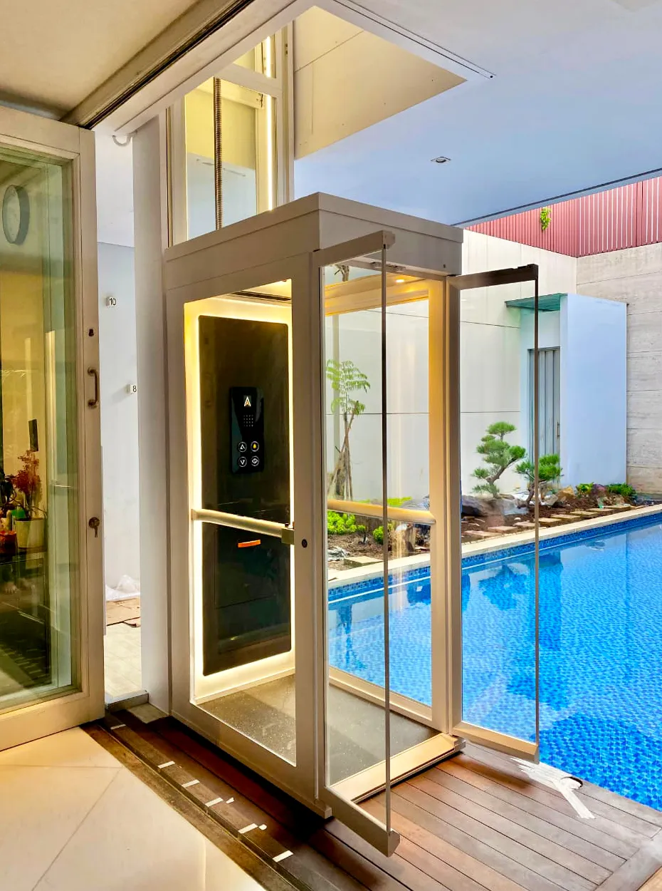 Lift rumah Ascenda by Cibes dengan desain kaca di area indoor dekat kolam renang, pencahayaan hangat dan akses praktis.